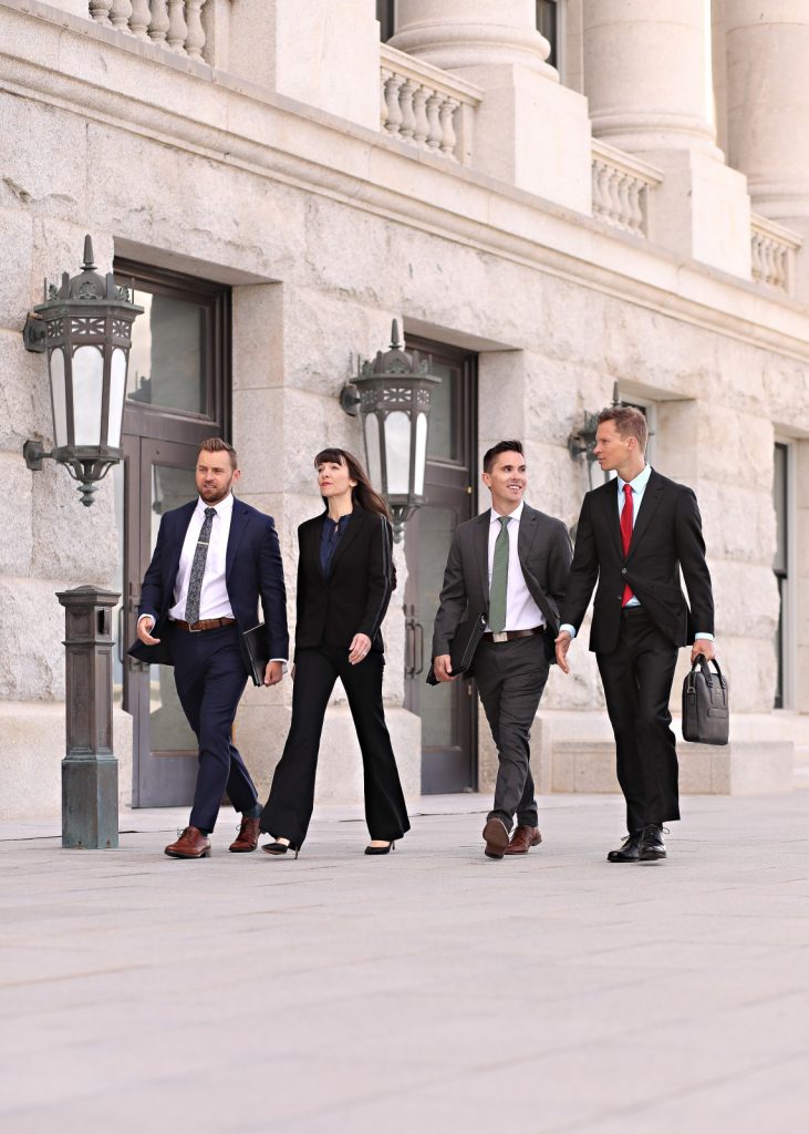 Four people in business attire walk together outside a stone building with large windows and ornate lanterns, appearing to be engaged in conversation.